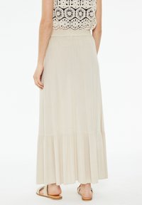 Falda maxi beige con un fondo acampanado, que presenta una textura suave y pliegues sutiles. Combinada con sandalias planas, resaltando un look casual.