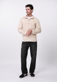 Smėlio spalvos megztinis su užtrauktuko kaklu ir krūtinės paneliu; derinamas su juodais susidėvėjusiais džinsais ir juodais sportbačiais su baltais dryžiais.