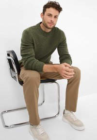 Grüner gestrickter Pullover mit gerippten Bündchen, beige Chinos und hellen Sneakers, die auf einem schwarzen und verchromten Stuhl vor einem weißen Hintergrund sitzen.