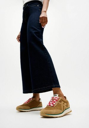 Persoon die donkere wijde jeans en beige sneakers met rode en witte veters draagt, staand tegen een effen witte achtergrond.