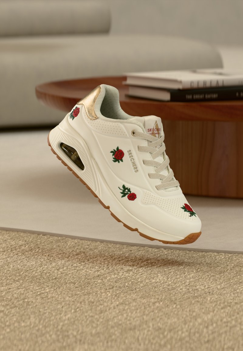 Biały trampek z czerwonym haftem róż, beżowymi sznurowadłami i przezroczystą podeszwą z poduszką powietrzną, unoszący się nad teksturowanym dywanem obok drewnianego stołu.