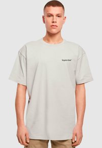 Camiseta de algodón de color gris claro y corte oversize, con un pequeño logotipo negro en el lado izquierdo del pecho. Mangas cortas, cuello redondo y ajuste relajado.