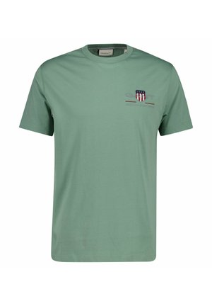 Grünes T-Shirt mit kurzen Ärmeln und Rundhalsausschnitt, mit einem kleinen roten, weißen und blauen Emblem und "1949" auf der linken Brust.