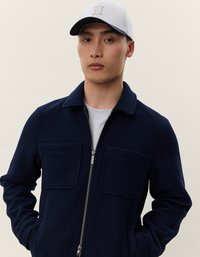 Young man wearing a navy blue zip-up jacket with front pockets and a white baseball cap with a beige Roman numeral II.