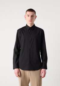 Camicia nera con bottoni, maniche lunghe, colletto, tessuto liscio, con un logo discreto sul petto. Vestibilità regolare e bilanciata.
