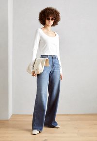 Blusa blanca de manga larga, jeans de tiro alto de pernera ancha azul, bolso beige y zapatos de color claro, sobre un fondo neutro.