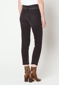 Femme portant un jean noir taille haute avec une taille cloutée et des fentes boutonnées aux chevilles, associé à des bottines marron en cuir de serpent.