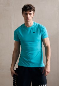 Türkisfarbenes sportliches T-Shirt mit kurzen Ärmeln und schwarzem Nike-Logo, figurbetontes Design, glatte Textur, getragen mit dunklen Shorts, Seil in der Hand gehalten.