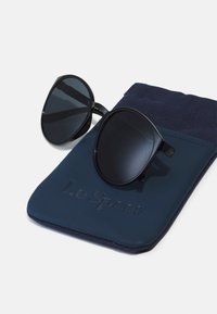 Černé sluneční brýle s kulatými skly, položené na tmavě modré tašce s embosovaným textem "Le Specs" a texturovaným prvkem z látky.