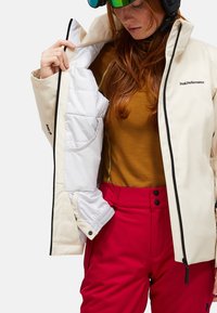 Kvindelig skiløber iført skitøj med en åben cremefarvet jakke, der viser en hvid polstret foring, en sennepsgul skjorte, røde bukser, skihjelm og reflekterende briller.