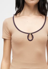 Γυναίκα που φοράει μπεζ ριμπ κοντομάνικο τοπ με σκουρόχρωμη επένδυση στο λαιμό και μικρή σχισμή σε σχήμα σταγόνας που κλείνει με χρυσό κουμπί στο κέντρο.