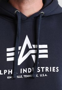 Marineblauwe hoodie met wit logo en tekst, voorzien van een trekkoordcapuchon en textuurstof. Het logo bevat een gestileerd sterrenpatroon.