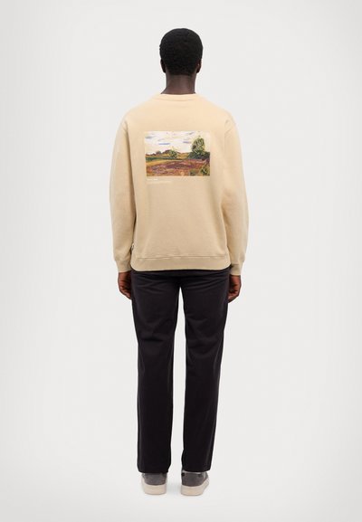 Béžová mikina s obdélníkovým krajinovým grafickým vzorem na zádech. Hladké manžety a lem, kombinováno s tmavými kalhotami a šedými botami.