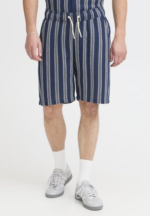 Navy blue striped shorts with elastic waistband and white drawstring. Made of textured fabric, reaching mid-thigh, paired with gray sneakers.