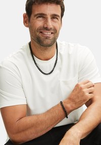 Zwarte ketting en armband gemaakt van gestructureerde schakels, gedragen met een witte T-shirt met ronde hals, model poseert met een ontspannen uitdrukking.