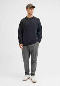 Dunkelgrauer Strickpullover mit einem gerippten Kragen, kombiniert mit hellgrauen Hosen und weißen Sneakers. Der gesamte Look ist lässig und entspannt.