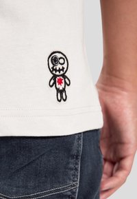 Camiseta de algodón blanca con un diseño bordado de un personaje de dibujos animados que presenta un patrón de calaveras y un detalle de corazón rojo cerca del dobladillo.