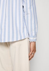 Camisa a rayas azul claro y blanca con puños abotonados, fabricada en algodón suave, con dobladillo curvado y corte relajado. Presentada con pantalones beige.