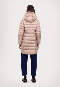 Mujer de pie de espaldas, vistiendo un abrigo largo acolchado beige con capucha, pantalones azul oscuro y botas negras, sobre un fondo blanco.