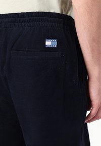 Marineblå corduroy joggebukser med elastisk midje, med en baklomme og en liten merkeetikett i blått og hvitt på høyre side.