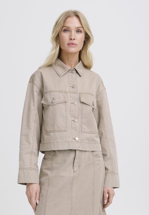 Donna bionda che indossa una giacca di jeans beige corta con tasche con bottoni e gonna a vita alta abbinata su uno sfondo semplice.