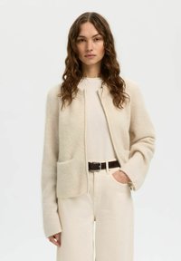 Femme aux longs cheveux bruns bouclés portant un cardigan à fermeture éclair beige clair, un haut crème, un pantalon beige clair et une ceinture marron foncé.