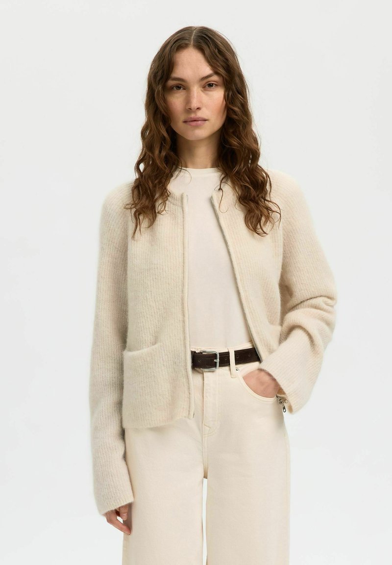 Femme aux longs cheveux bruns bouclés portant un cardigan à fermeture éclair beige clair, un haut crème, un pantalon beige clair et une ceinture marron foncé.