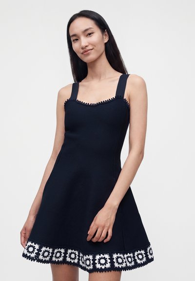 Jeune femme aux longs cheveux foncés portant une robe bleu marine sans manches avec de larges bretelles et un ourlet blanc à motif floral.