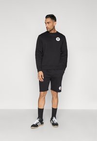 Melns džemperis un šorti, kokvilnas maisījums. Džempera priekšpusē ir apaļas formas logo uzšuve; šortiem ir līdzīga uzšuve. Valkāts ar melnām zeķēm un kurpēm.