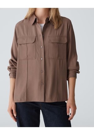 Woman wearing a long-sleeve brown button-up shirt with two chest pockets and dark blue pants, standing against a plain background.