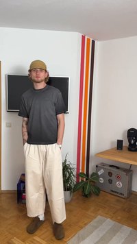 Pilkos trumpomis rankovėmis marškiniai, laisvi kreminiai kelnes ir rudi klumpiai. Šviesus sienų dekoro elementas su spalvingomis vertikaliomis juostomis. Medinis grindys ir minimalūs baldai.