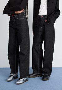 Black high-waisted wide-leg denim jeans with contrasting white stitching and a red accent button, paired with grey and black footwear.
