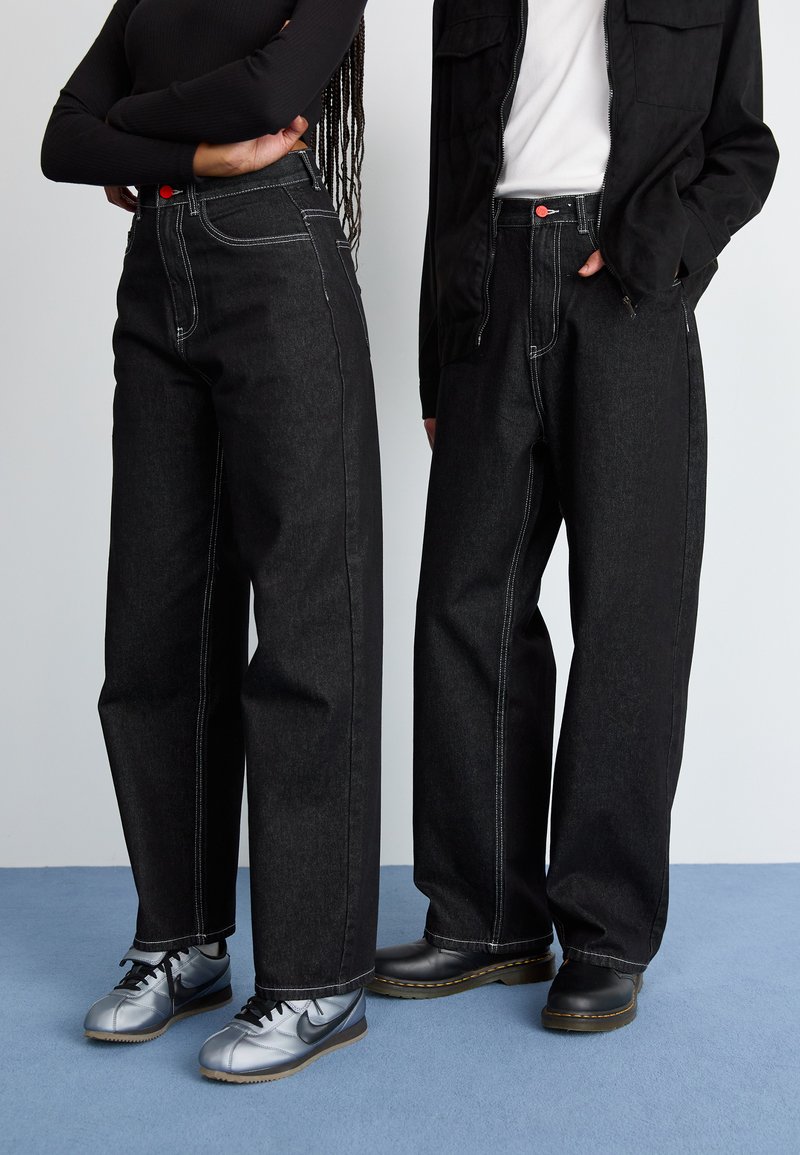 Black high-waisted wide-leg denim jeans with contrasting white stitching and a red accent button, paired with grey and black footwear.