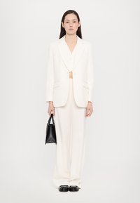 Blazer blanc cintré sur un crop top blanc et un pantalon large, le tout en tissu texturé. Tenant un petit sac à main noir. Chaussures noires.