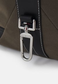 Sac vert olive avec des accents noirs, doté d'un fermoir en métal argenté de type mousqueton et de détails de surpiqûre contrastants. Matière en tissu texturé.