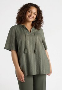 Blusa verde oliva con mangas cortas, tela texturizada, rayas verticales y dos borlas en el escote. Combina con pantalones a juego.