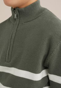 Stickad grön tröja med halvdragkedja, med en ribbad halsringning och vita horisontella ränder över bröstet. Texturerad tyg.