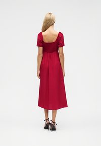 Γυναίκα με μακριά ξανθά μαλλιά που φοράει κόκκινο μίντι φόρεμα με φουσκωτά μανίκια και μαύρα ψηλοτάκουνα σανδάλια, στέκεται με την πλάτη προς τα εμπρός.