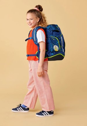 Der Rucksack hat einen blauen Stoff mit grünen Akzenten und Blitzmustern, getragen über einem roten Pullover und gestreiften Hosen, dazu marineblaue Sneakers.