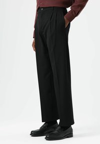 Pantalon noir plissé à coupe droite, confectionné dans un tissu lisse. Porté avec des mocassins noirs et une chemise à manches longues.