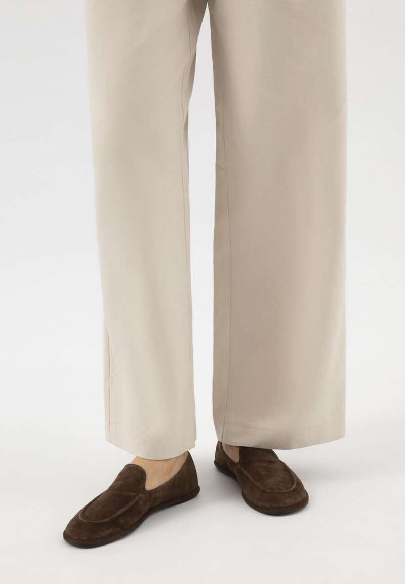 Pantaloni beige a gamba larga indossati con mocassini scamosciati marrone scuro su sfondo bianco.