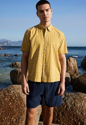 Man wearing short-sleeve yellow patterned button-up shirt and navy shorts standing on rocky shore with ocean and mountains in background.