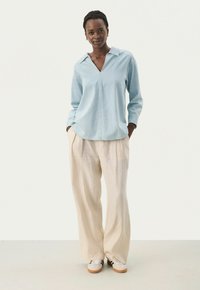 Camicia azzurra a maniche lunghe con colletto e mezza patta, abbinata a pantaloni ampi beige chiaro e sneakers bianche. Texture lisce.