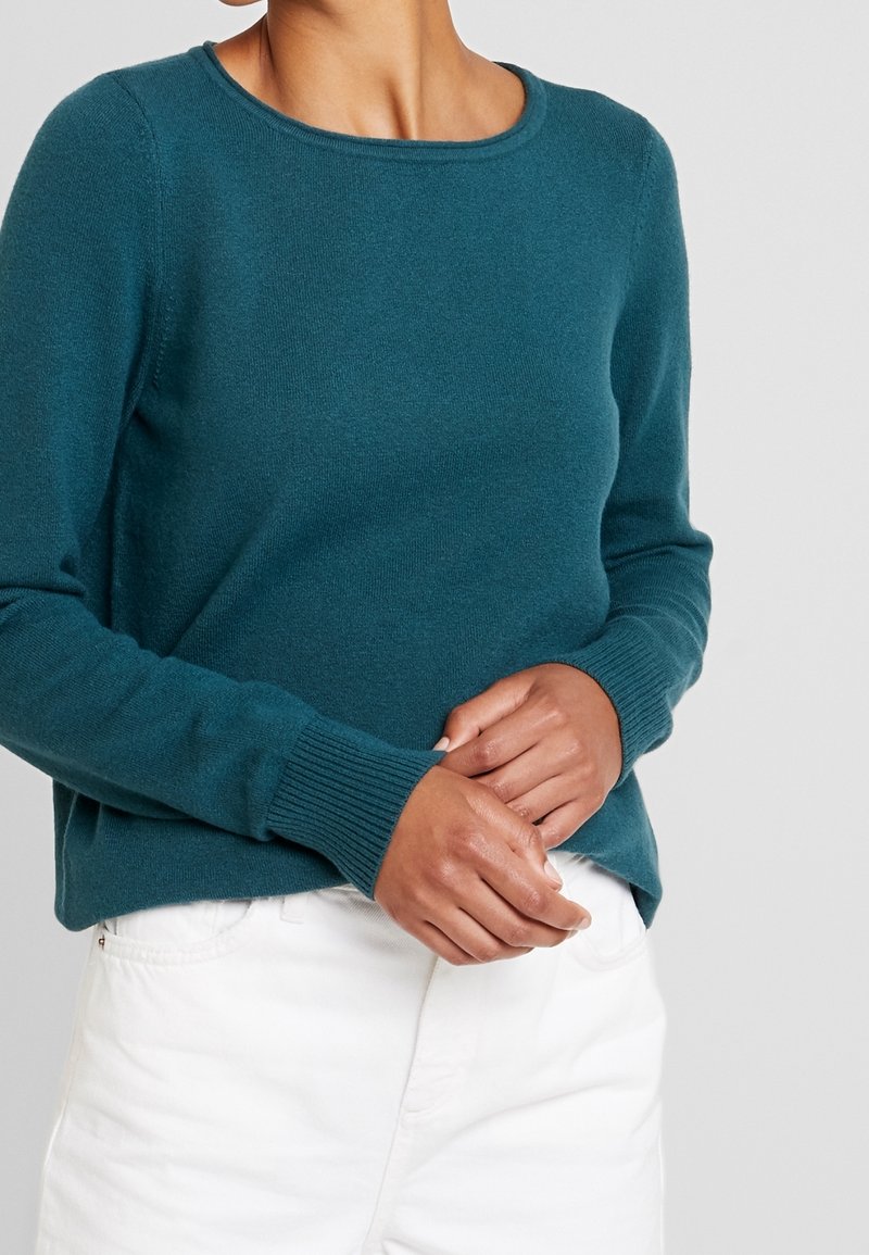 Personne portant un pull en maille manches longues couleur sarcelle et un pantalon blanc, mains doucement jointes à la taille devant un fond uni.