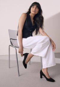 Chaleco negro sin mangas con botones, pantalones anchos blancos y zapatos de tacón alto de ante negros. Silla transparente con patas de cromo. Fondo neutro.