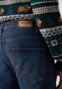 Gebreide trui met een bos thema in groen, rood en beige, gecombineerd met donkerblauwe denim jeans met een uniek ontwerp van de achterzak.