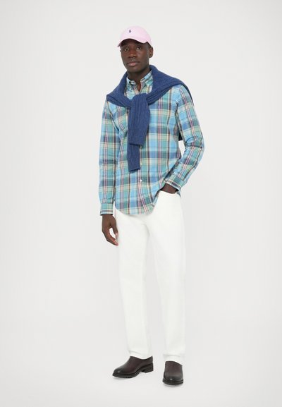 Karirasta srajca v modri, zeleni in roza barvi, z mornarsko modrim puloverjem, vezanim okoli vratu, belimi hlačami in črnimi čevlji. Rozasta kapa zaokroži videz.