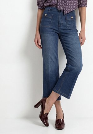 Frau trägt blaue verkürzte Jeans mit Fronttaschen und braune Loafers mit hohen Absätzen und steht vor einer weißen Wand.