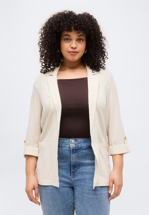 Kvinne med krøllete hår iført beige åpenblazer, mørkebrun topp og høytlivsblå jeans som står mot en ensfarget bakgrunn.