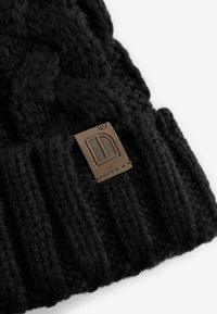 Bonnet en tricot noir avec un motif en câble et une bande côtelée. Présente une étiquette en cuir marron avec un logo, mettant en valeur le tissu texturé.